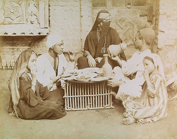 Arab family photographed during dinner. The members of the small nucleus are seated on the ground around a rudimentary table on which is placed a tray filled with dishes of food
